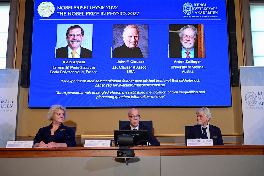 Three middle-aged people sit at a conference table in front of a screen displaying the winners of the Nobel Prize in Physics.