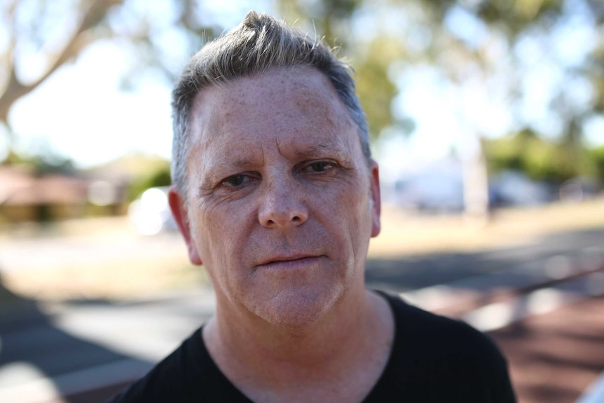 A headshot of a man with a black t-shirt standing on a street.