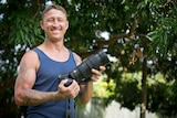 A man in a blue singlet holds a large camera in his hands while standing in a leafy back garden.