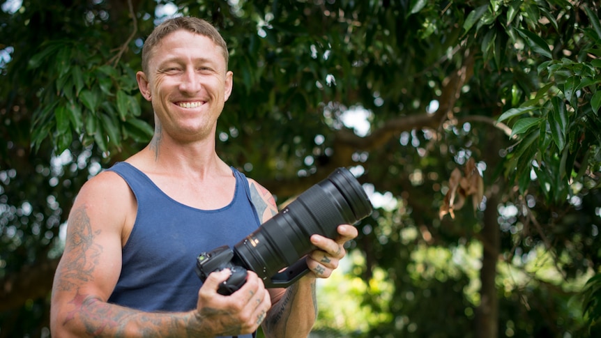 A man in a blue singlet holds a large camera in his hands while standing in a leafy back garden.