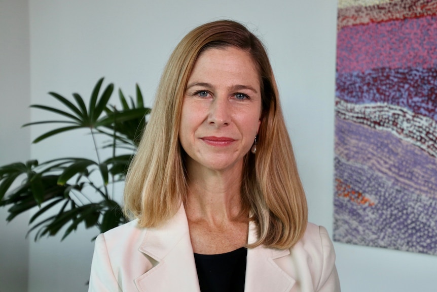 A woman with should length dark blond hair wearing a white suit jacket smiles into the camera