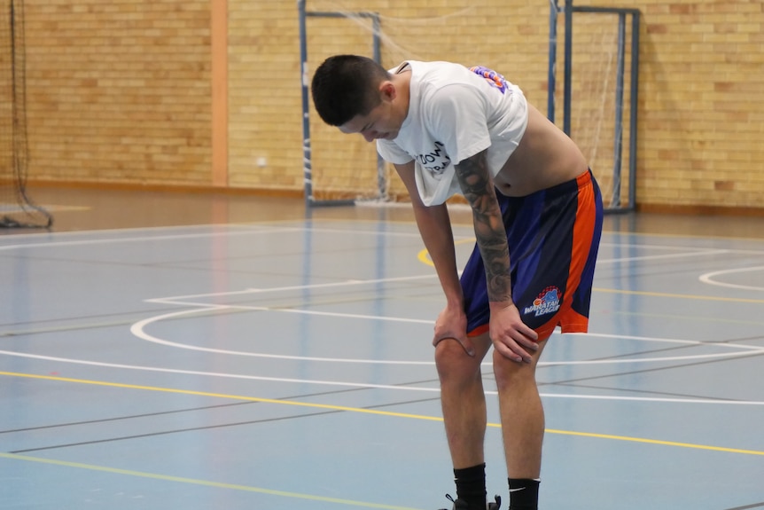 A young basketballer has his hands on his knees and is hunched over, exhausted.