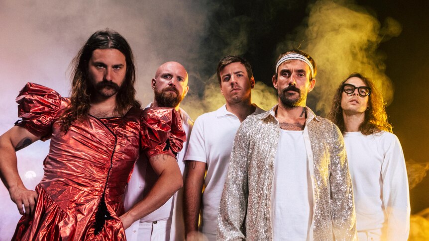 Five men from British band IDLES stand together looking at the camera. One man wears a sparkly red dress