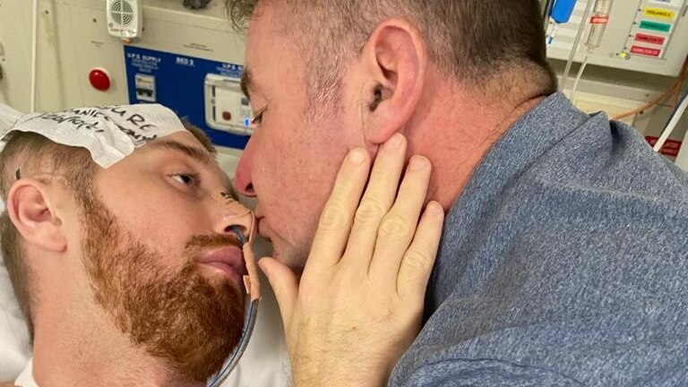 Danny Hodgson's father leans in to give his son a kiss while he is lying in a hospital bed.