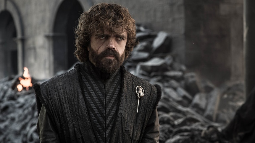 Actor Peter Dinklage, wearing leather gillet with pin of hand, stands looking fed up, with building ruins/rubble behind him.