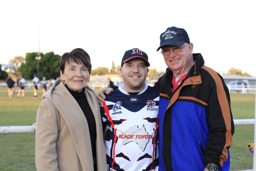 A young man in a footy jersey stands between his parents on a footy field.