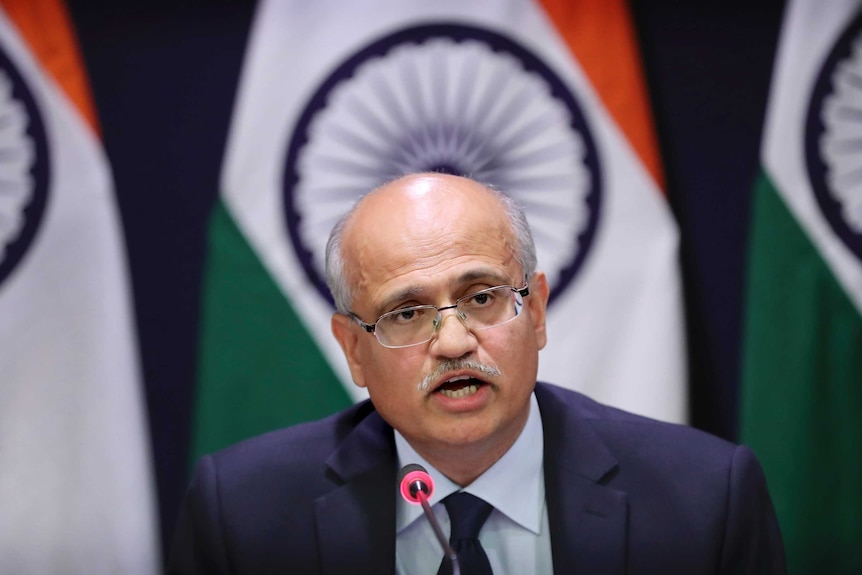 Vijay Gokhale wears a navy suit and clear glasses. He is mid-speech, has a microphone in front of him and is in front of flags.