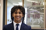 A boy wearing a tie and blazer stands in front of a window that has "be kind" painted on it