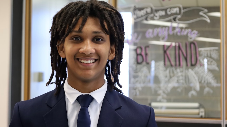 A boy wearing a tie and blazer stands in front of a window that has "be kind" painted on it