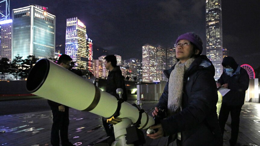 A woman looks into the sky as she touches a big telescope.