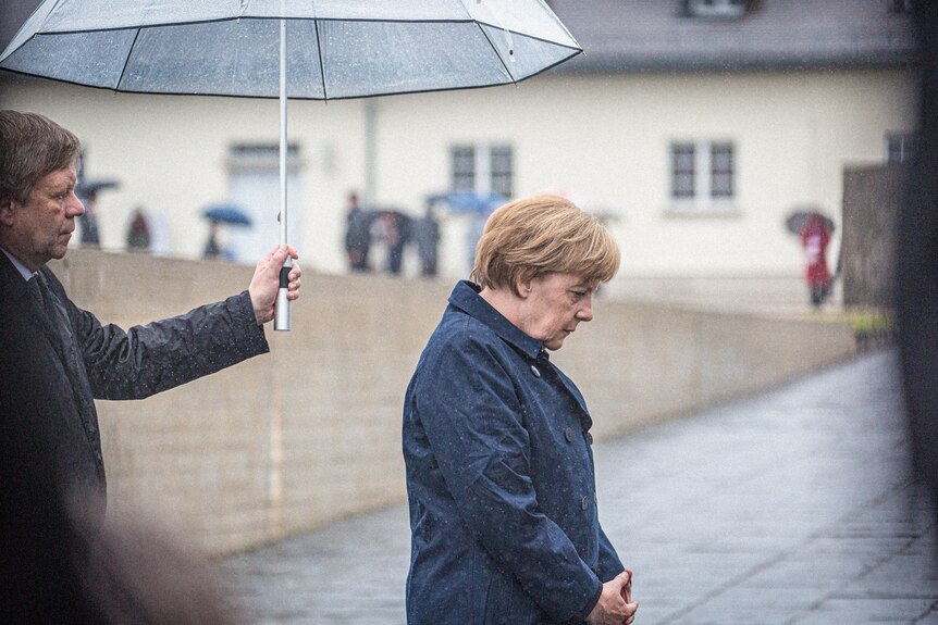 Angela Merkel wearing a dark coat looks down on a rainyy day as a man holds an umbrella over her.