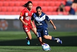 A soccer player wearing dark blue dribbles the ball away from a player wearing red