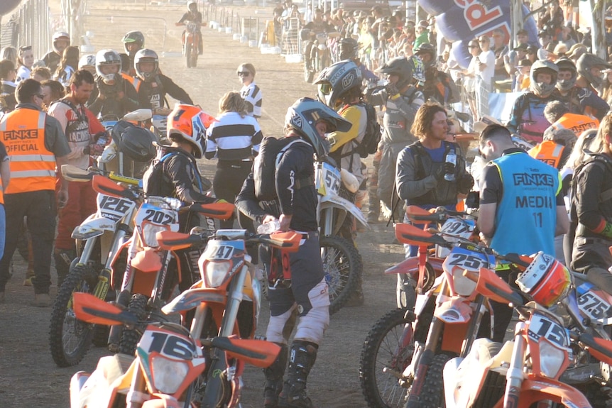 A crowd of dirt bike riders in protective gear gathered on a road in the outback.