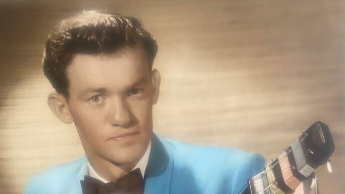 Grainy colour photo of young man smiling, holding guitar, and wearing bright blue suit and bow tie.