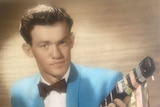 Grainy colour photo of young man smiling, holding guitar, and wearing bright blue suit and bow tie.