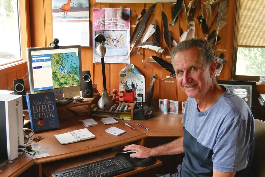 Allan Briggs sits at his desk, near his computer and bird guide with feathers and a bird calendar on the wall.