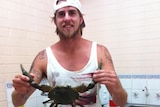 Ben Gerring stands smiling holding a mud crab by its claws in a kitchen, wearing a white tank top and a white hat backwards.