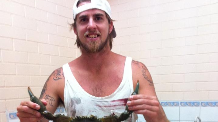 Ben Gerring stands smiling holding a mud crab by its claws in a kitchen, wearing a white tank top and a white hat backwards.