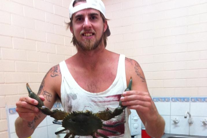 Ben Gerring stands smiling holding a mud crab by its claws in a kitchen, wearing a white tank top and a white hat backwards.