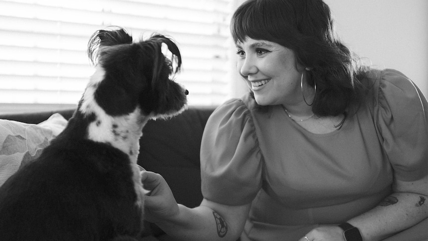 A smiling woman with shoulder length hair and a small black dog look at each other while sitting on a couch together 