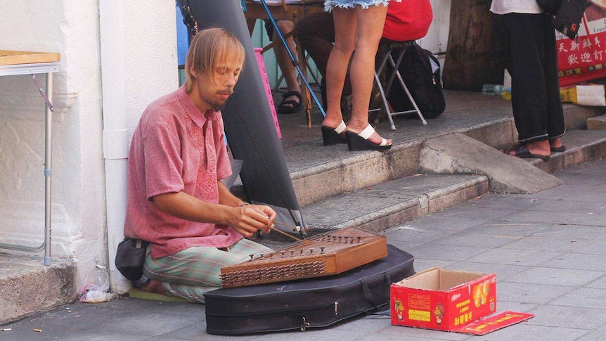 Western begpacker plays an Asian musical instrument on the street.