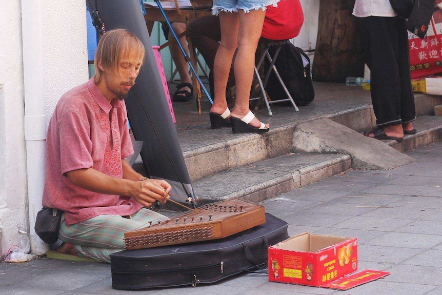 Western begpacker plays an Asian musical instrument on the street.