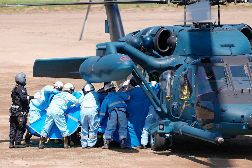 Emergency workers in white overalls hold large blue privacy screens near the door of a large helicopter.