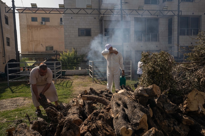 A man covers his eyes as smoke billows from a firepit, while surrounded by piles of wood 