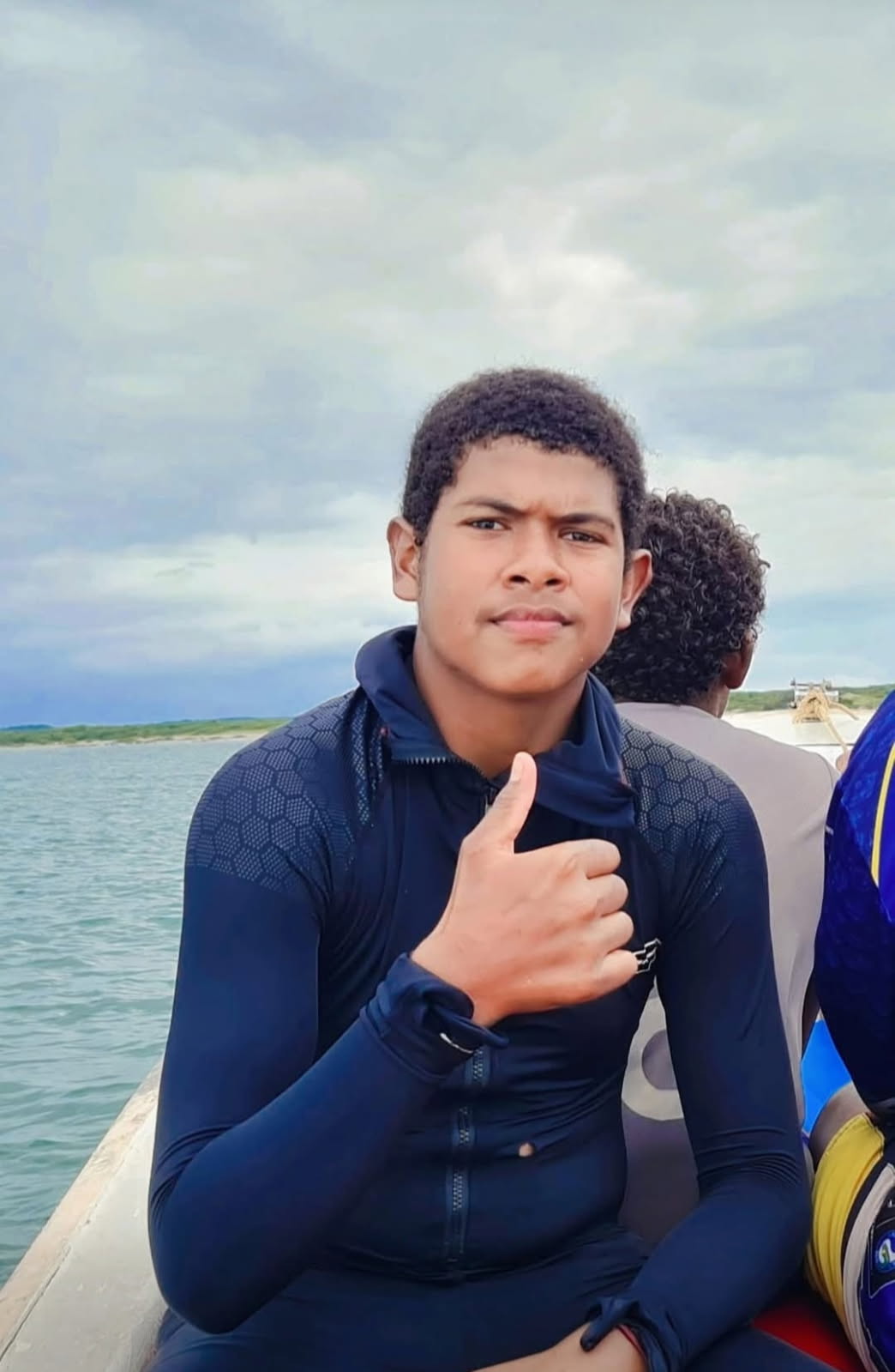 A young man with short, dark hair gives the thumbs up as he sits with others on a small boat beneath a cloudy sky.
