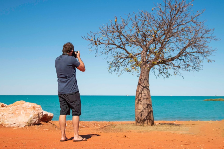Man taking photo of boab tree, with ocean in background