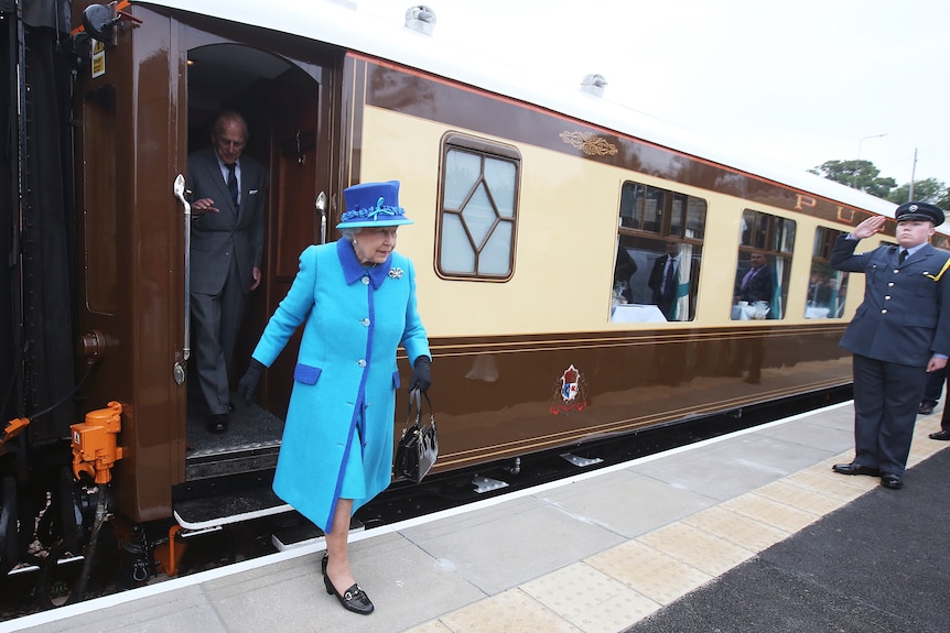 Queen Ellizabeth is wearing a blue outfit as she steps off the royal train and onto a platform. 