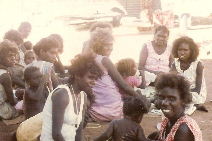 A group of Indigenous women and children sit on the ground. They are all smiling to the camera and are happy.