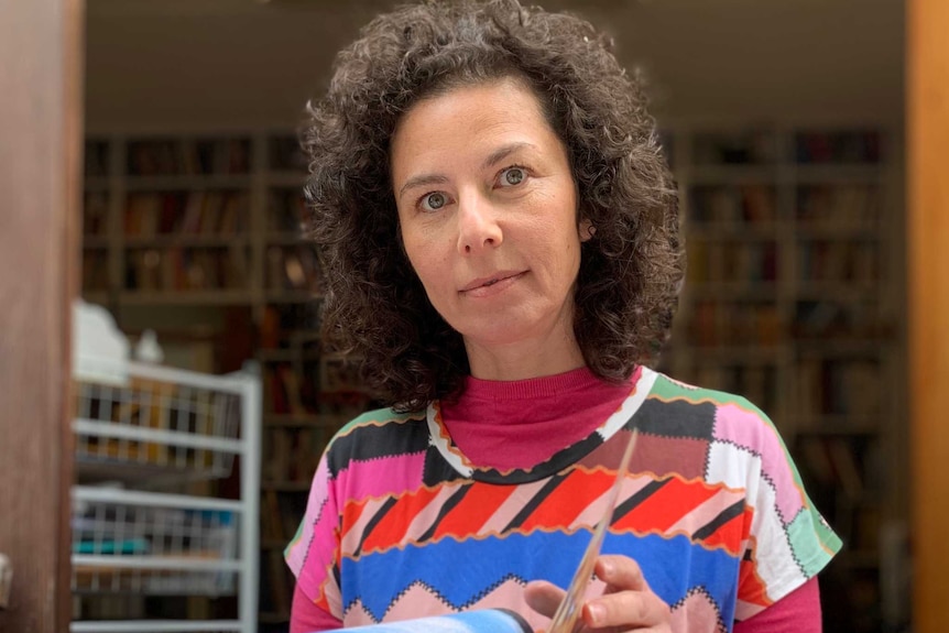 a woman in a multi-coloured shirt opens the pages of a book while looking at the camera