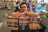 Children's author Oliver Phommavanh with his arms and head resting on a big pile of his books.