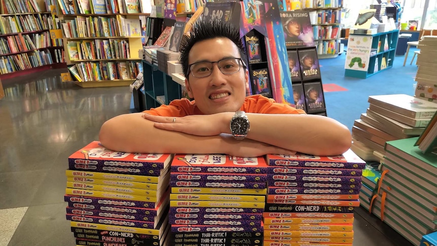 Children's author Oliver Phommavanh with his arms and head resting on a big pile of his books.