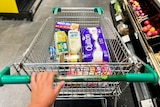 A Woolworths trolley filled with toliet paper, instant coffee, tampons, milk, eggs, bread and Weetbix.