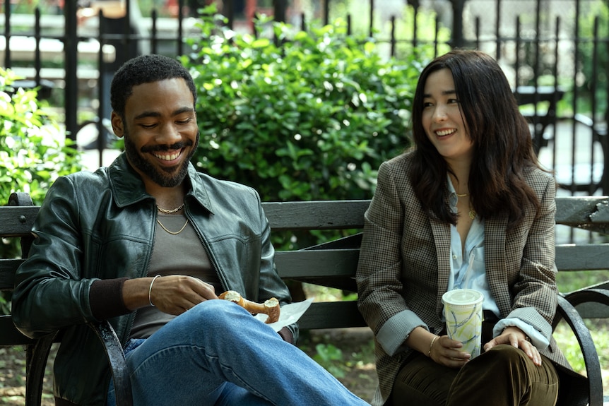 Donald Glover and Maya Erskine sitting on a park bench smiling, one holding a drink, both in character as Mr & Mrs Smith