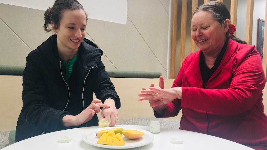 Two smiling woman applying moisturiser on their hands with an open mango on the desk in front of them.