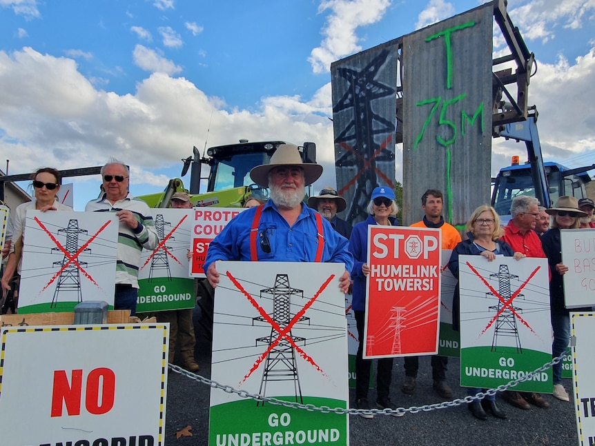 man stands with protest sign against overhead transmission lines for HumeLink project.