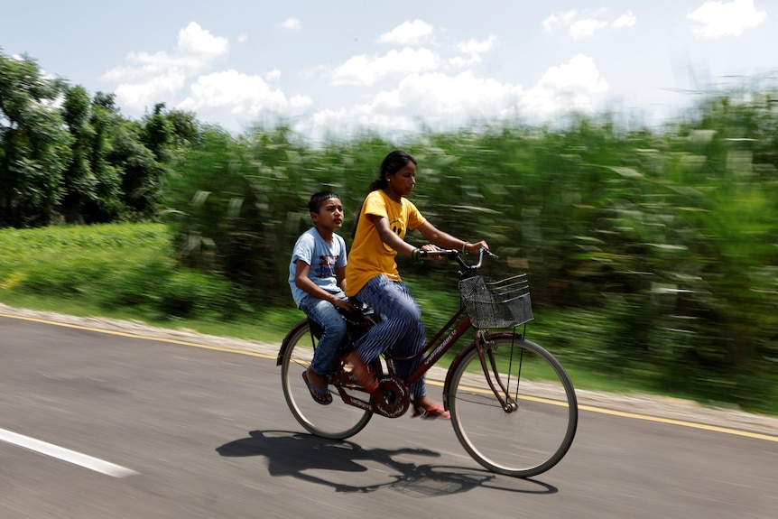 A woman and a young boy ride on a bicycle on a road as fields rush past them
