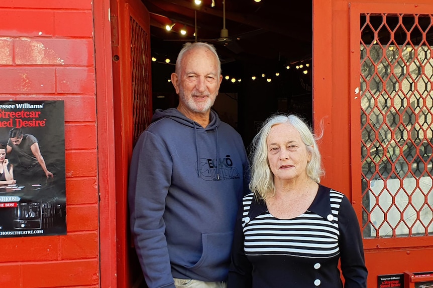 A man and a woman with white hair stand in front of a red door