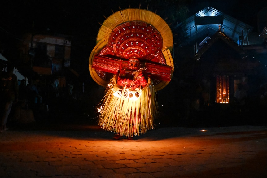 A person wears a costume of an Indian god with a grass skirt and large red headdress at a traditional dance.