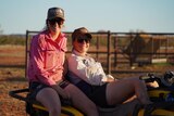 Two smiling young girls wearing a pink shirt, cap and glasses sit on a bike.