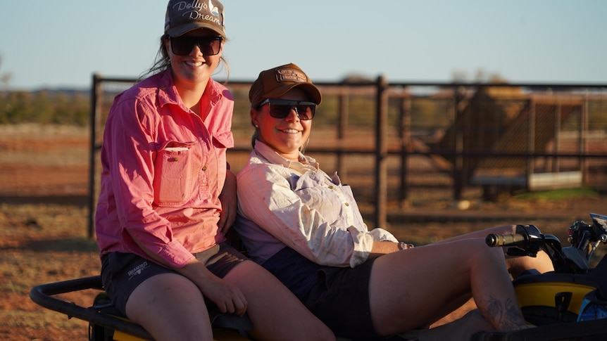 Two smiling young girls wearing a pink shirt, cap and glasses sit on a bike.