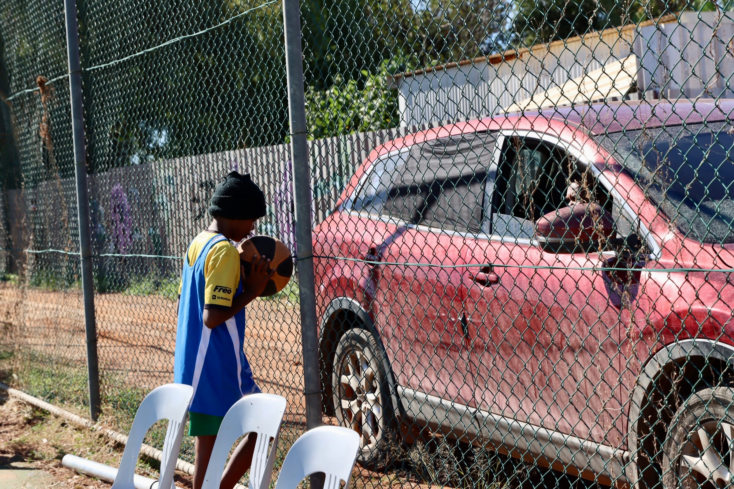 An Aboriginal boy holding a basketball looking through the fence at red car with window down