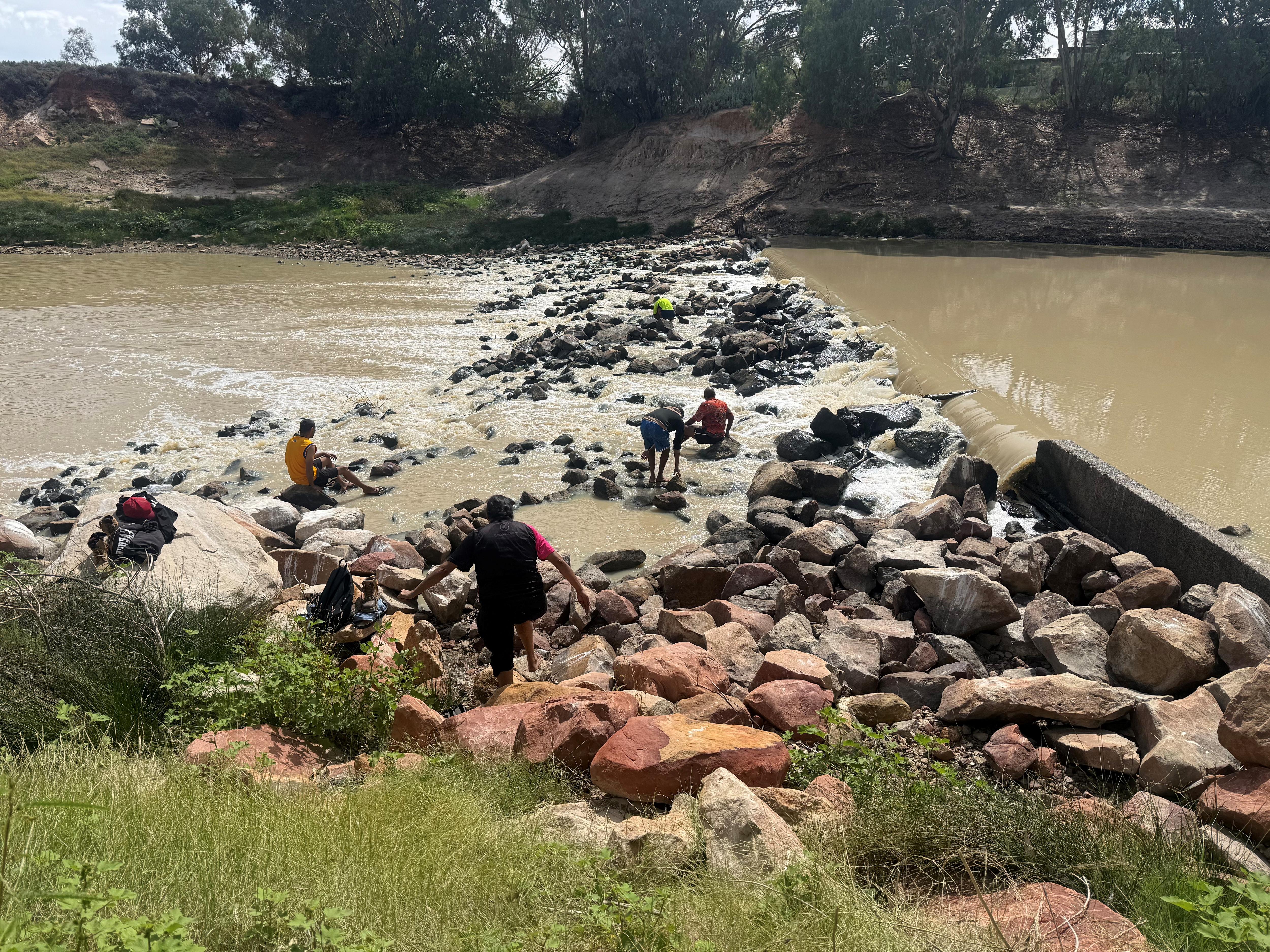 A landscape of a river with a rock weir connecting one bank to the other with five people climbing on the rocks