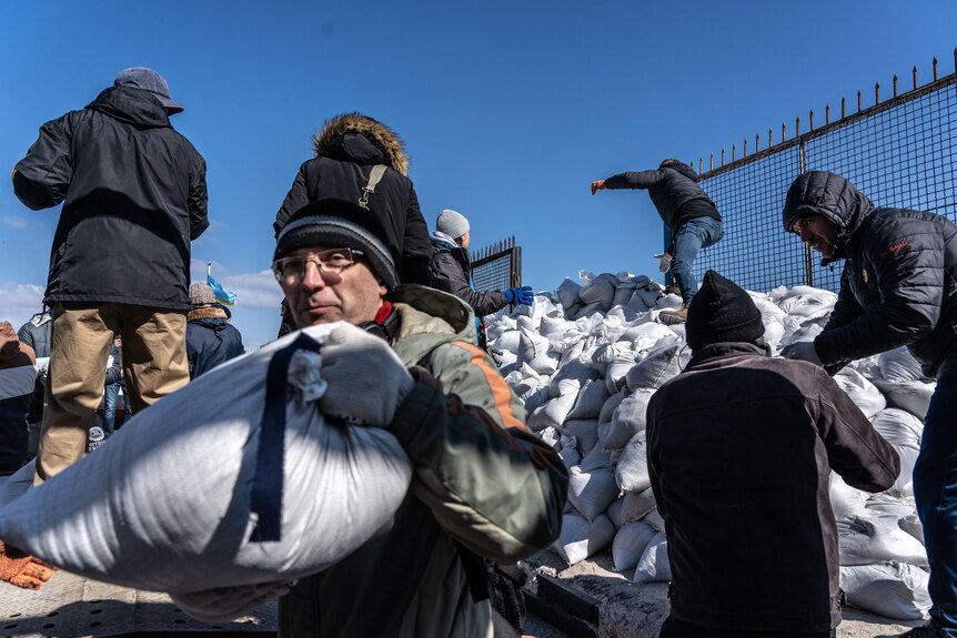 A man carries a sandbag away as other men climb over a pile of sandbags.