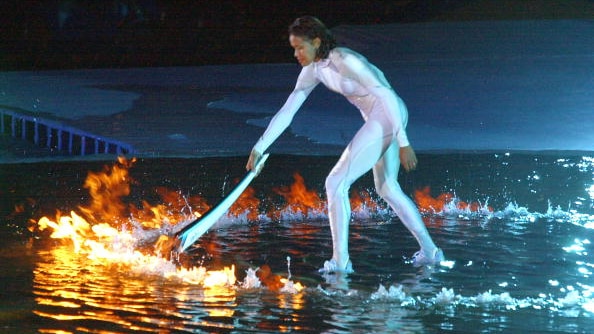 A woman stands in a pool of water, holding a flaming torch down to light another ring of fire at her feet