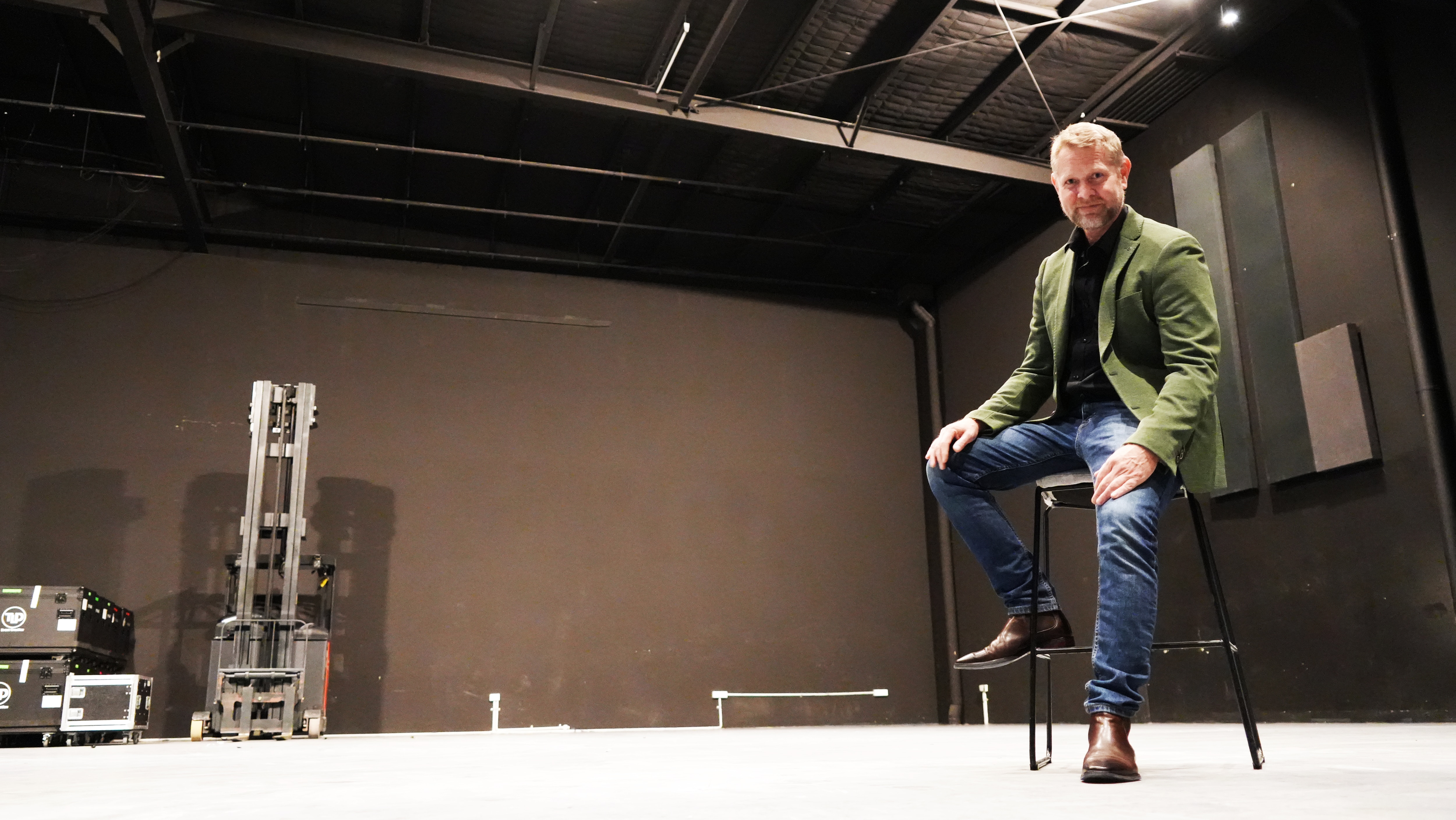 A smartly dressed man (Chris Amos) sitting on high stool smiling in a studio.