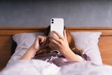 A woman lies in bed, under the covers, messaging on her phone.